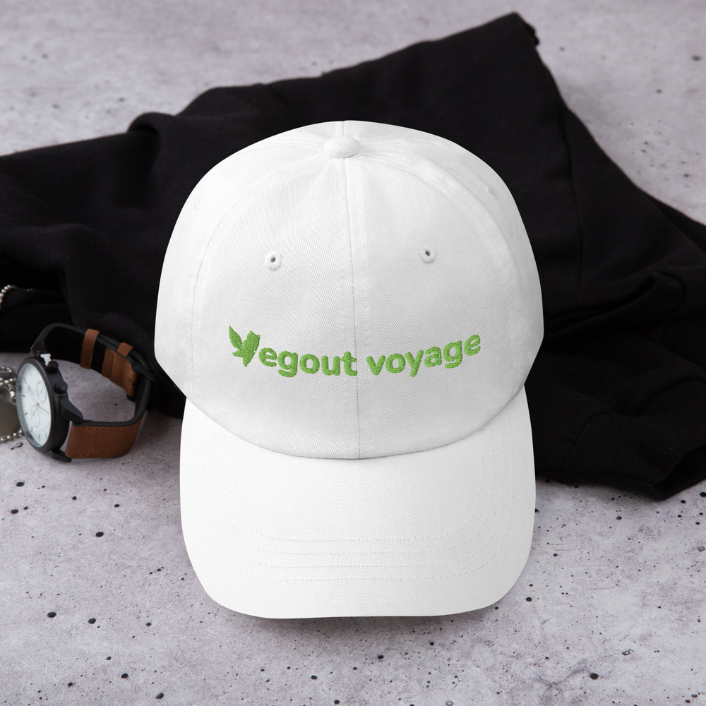 White cap with 'Vegout Voyage' logo on a gray surface with black fabric and watch.