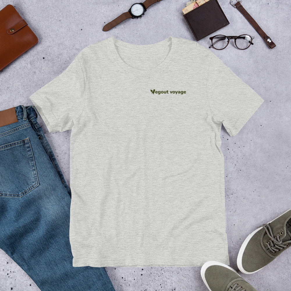 Flat lay of a light heather gray "Vegout Voyage" logo t-shirt paired with jeans, watch, glasses, and shoes.